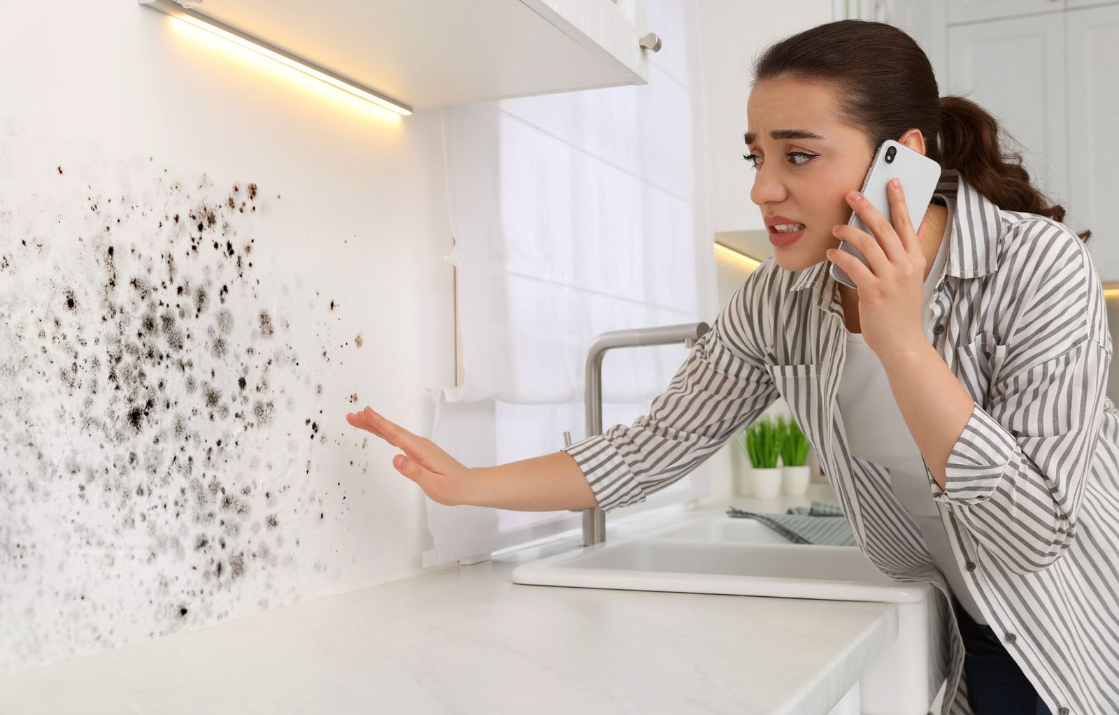A woman speaks on a cell phone in a kitchen with visible mold on the wall, highlighting the need for mold removal services.