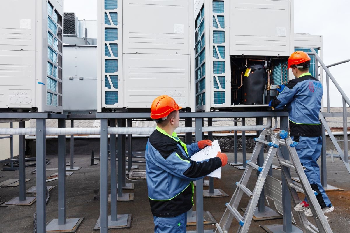 Two men repairing a large air conditioning unit, focused on their task in a well-lit mechanical room.
