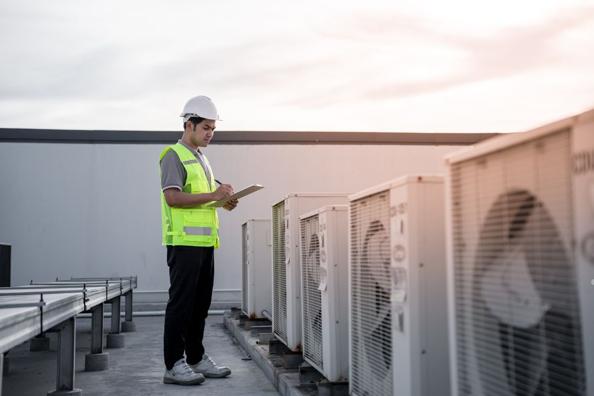 A man wearing a safety vest stands beside several air conditioning units in an outdoor setting.
